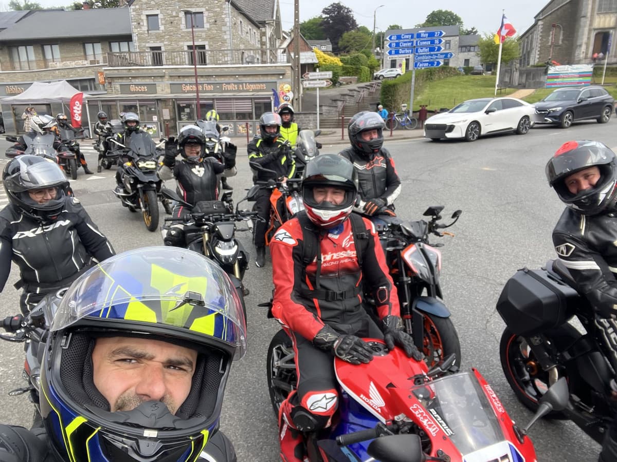 Een groep motorrijders met helm op en hun motoren op een pleintje in een dorp, met een lachende rijder vooraan in beeld.