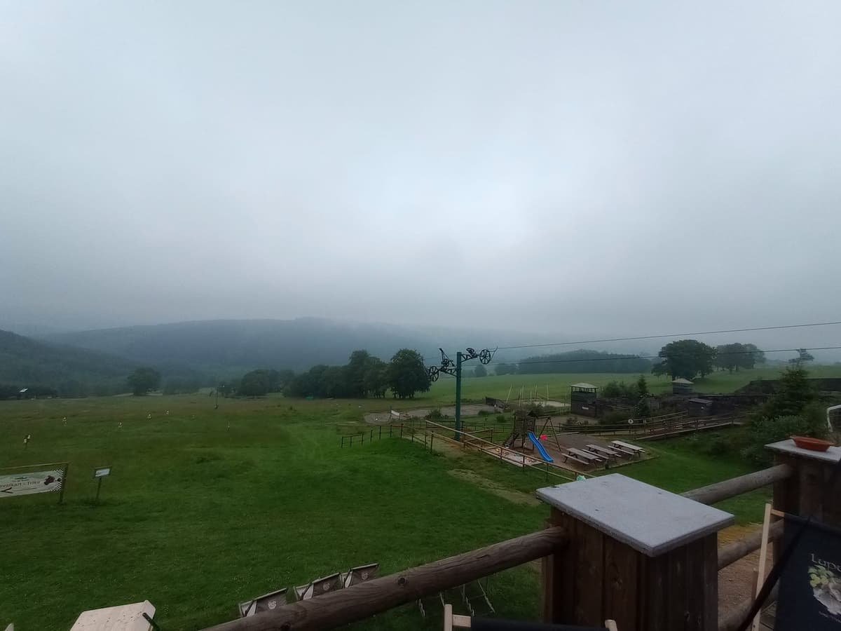 Uitzicht vanaf een houten terras over een groene helling met een skilift en heuvels in de mist.