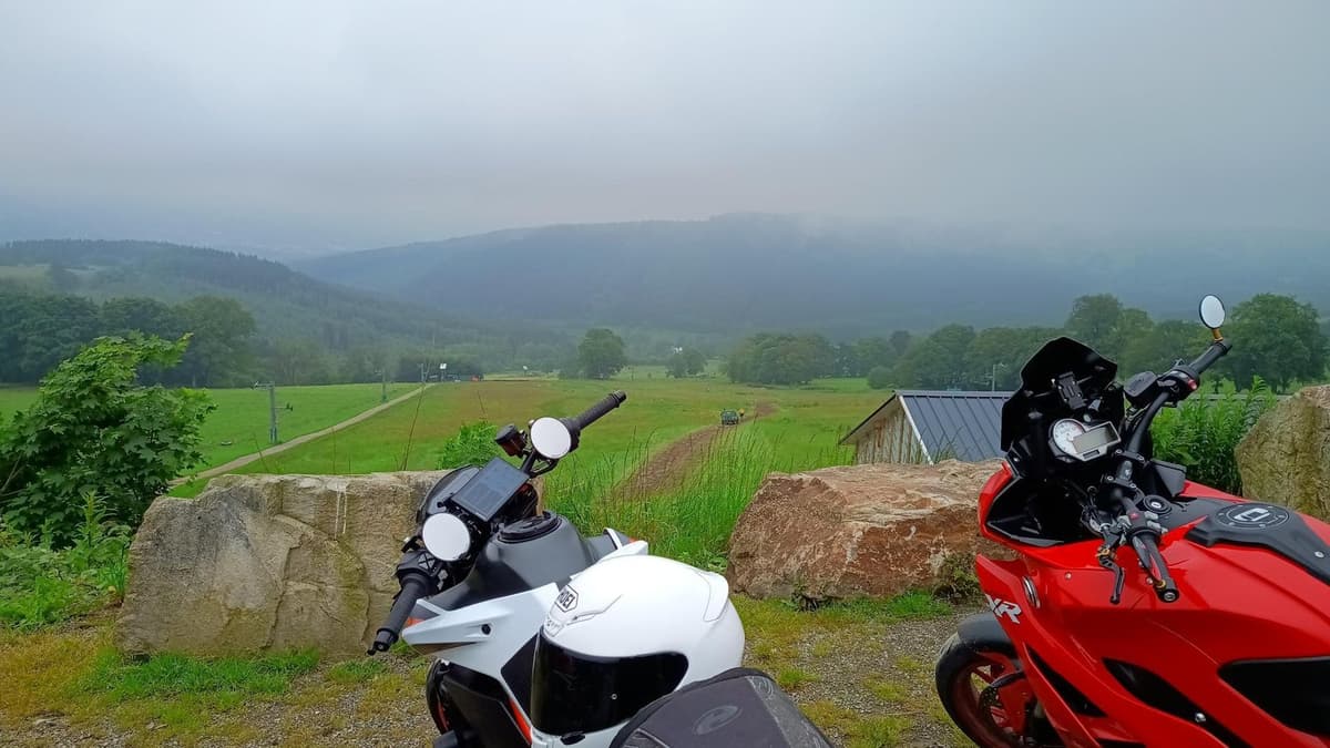 Een witte en een rode motor tussen grote rotsblokken, met daarachter groene weiden en beboste heuvels in de mist.