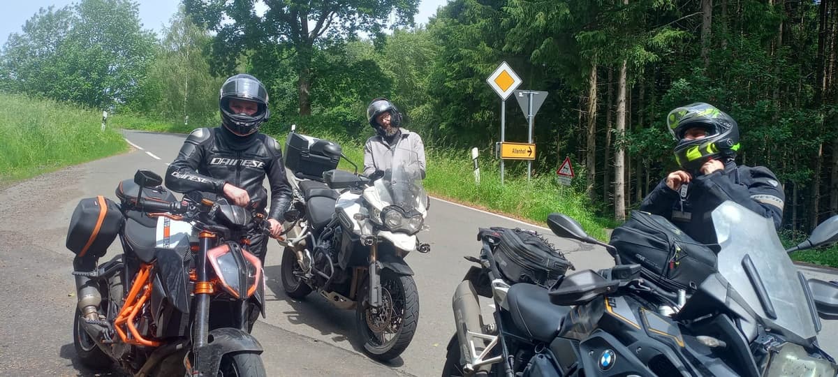 Drie motorrijders met helm op staan langs een landweg bij hun motoren, een oranje KTM, een witte Triumph en een zwarte BMW, aan een kruispunt in het groen.