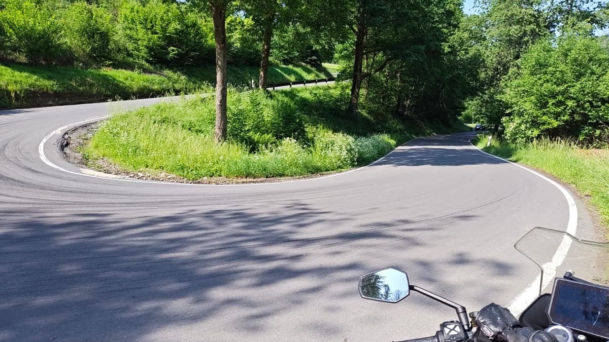 Zicht vanaf een motor op een haarspeldbocht in een smalle weg tussen groene bermen en bomen, met de spiegel en het dashboard vooraan.