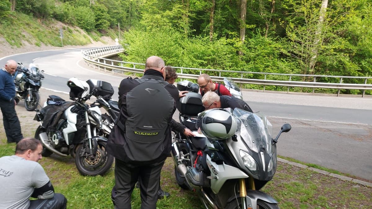 Motorrijders buigen zich over een kaart op een motor, in de berm van een bochtige weg langs het bos.