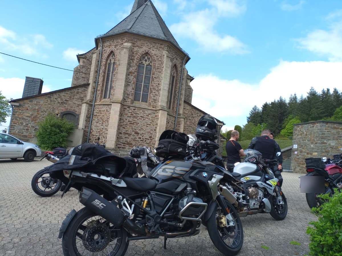 Een BMW GS en andere motoren op een pleintje voor een stenen kerk, met een paar motorrijders die staan te praten.