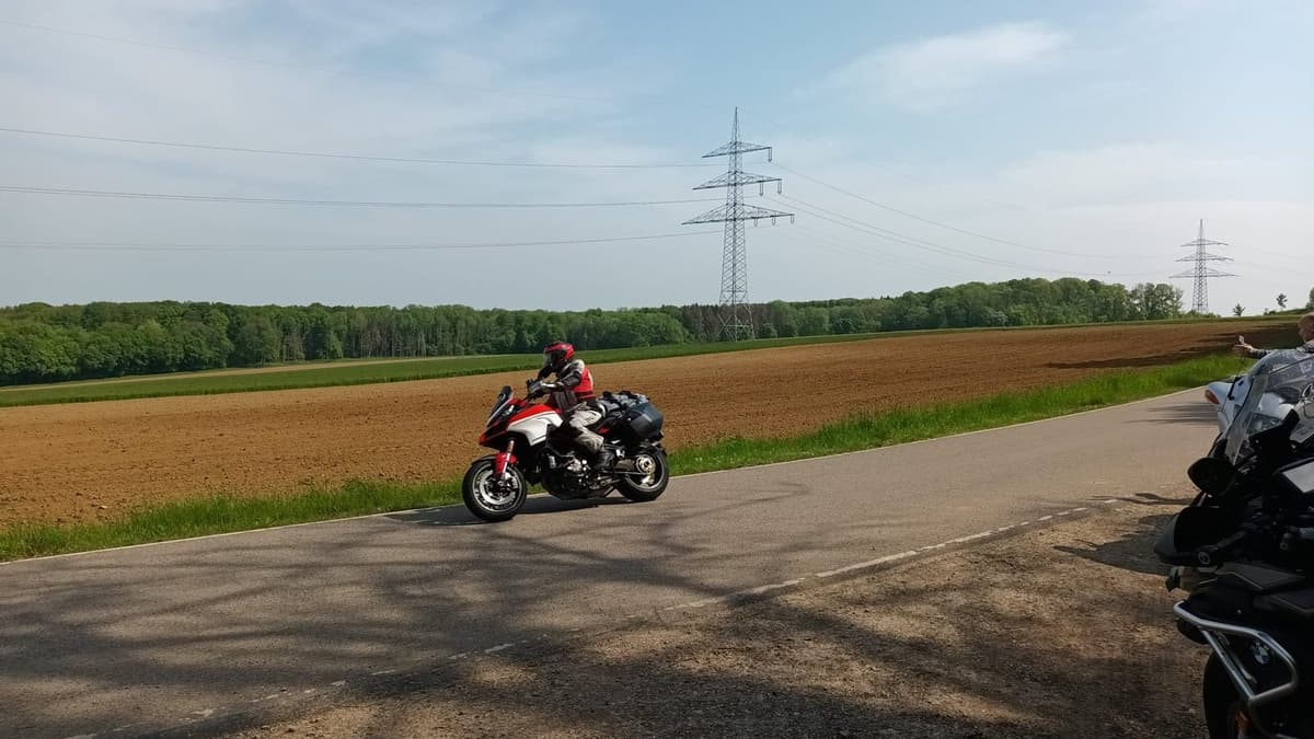 Een rood-witte motor met koffers rijdt over een landweg langs omgeploegde velden, met een bosrand en hoogspanningsmasten onder een blauwe lucht.