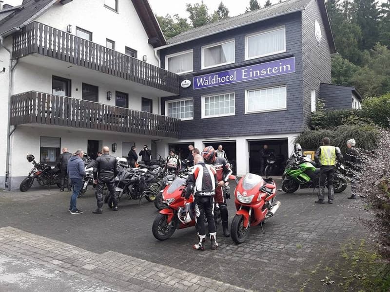 Motorrijders en hun motoren voor een hotel met houten balkons en het opschrift Waldhotel Einstein, tussen de bomen.