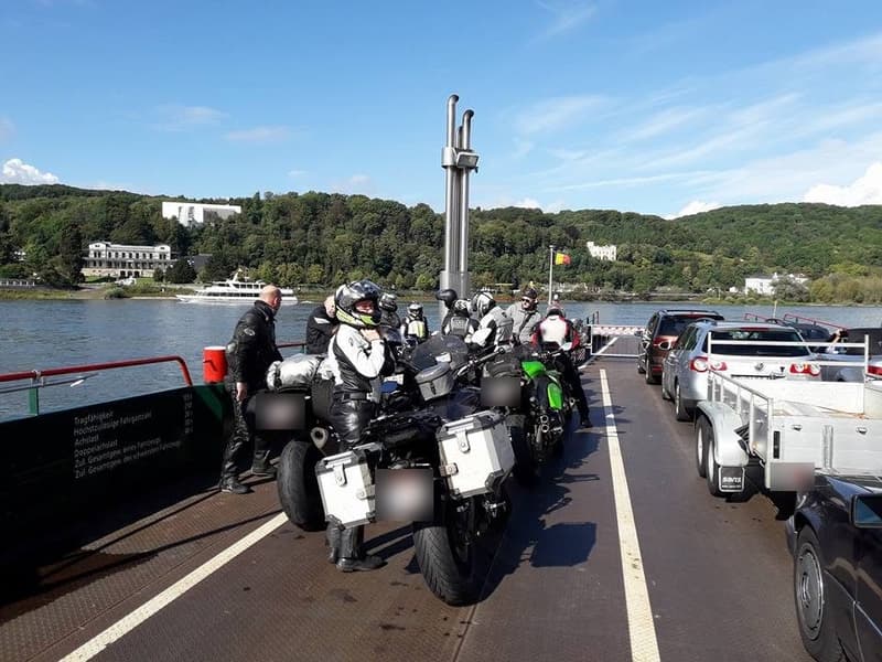 Motorrijders met hun motoren op een veerboot over de Rijn, met beboste heuvels en een rondvaartboot aan de overkant.