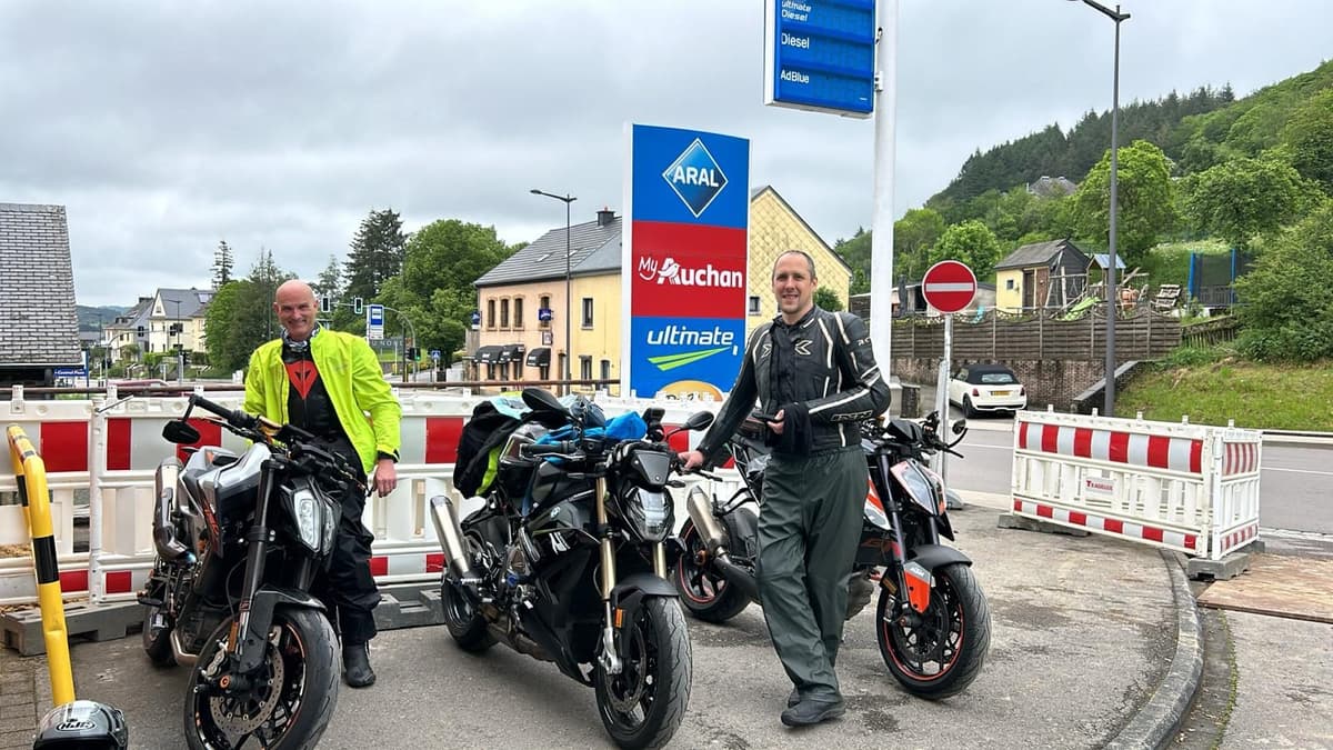 Twee lachende motorrijders bij drie naked bikes aan een tankstation in een dorp, tussen rood-witte werfhekken, met beboste hellingen op de achtergrond.