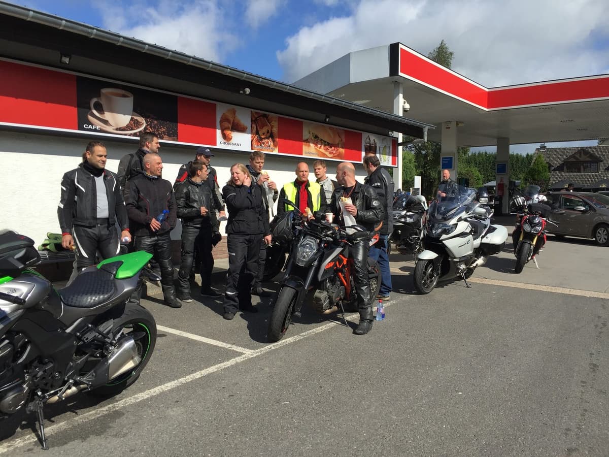 Een groep motorrijders staat te praten aan een tankstation onder een blauwe lucht, sommigen met een broodje of een flesje water, met vooraan een groene en een oranje naked bike en een witte BMW-toerfiets.