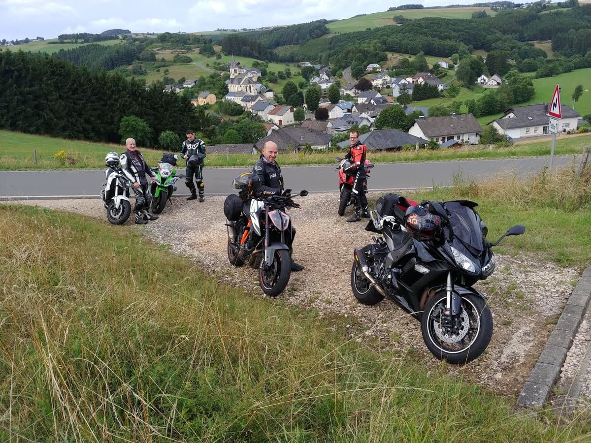 Motorrijders bij hun motoren op een grindstrook langs de weg, boven een dorp met een kerktoren in een groen, heuvelachtig landschap.