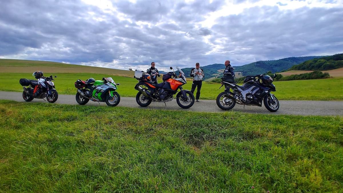 Vier motoren op een rij langs een smalle weg door groene weiden, met drie motorrijders erbij en beboste heuvels onder een grijze wolkenlucht.