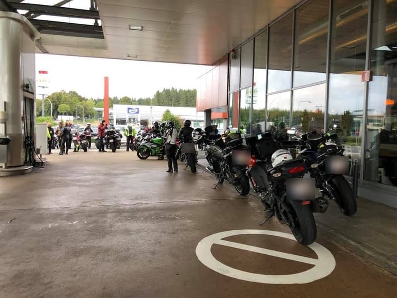 Een rij motoren onder de overkapping van een tankstation, met motorrijders in pak die verderop bij hun motoren staan te wachten.
