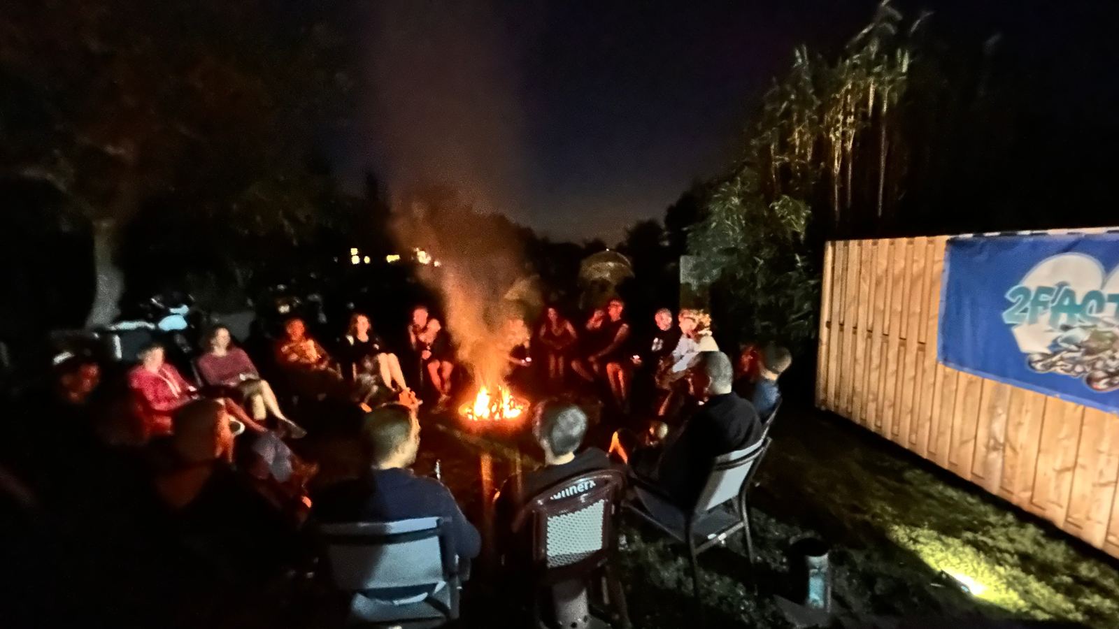 Een groep zit 's avonds in een kring rond een kampvuur in een tuin, naast een 2fast4u-spandoek.