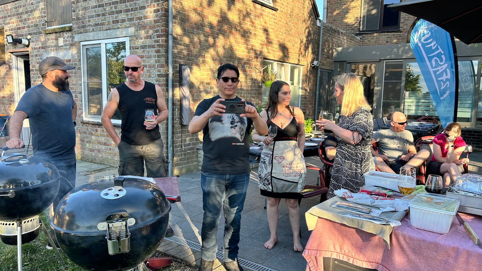 Een groep staat te praten rond twee barbecues op een terras in de avondzon.