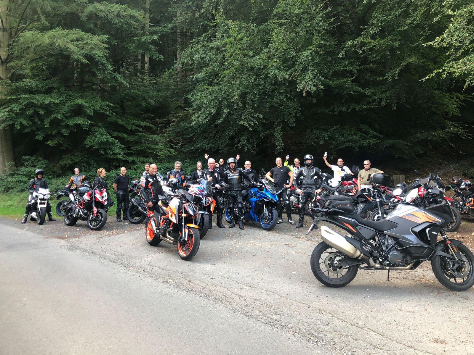Een grote groep motorrijders staat lachend en zwaaiend bij hun motoren langs een bosweg.