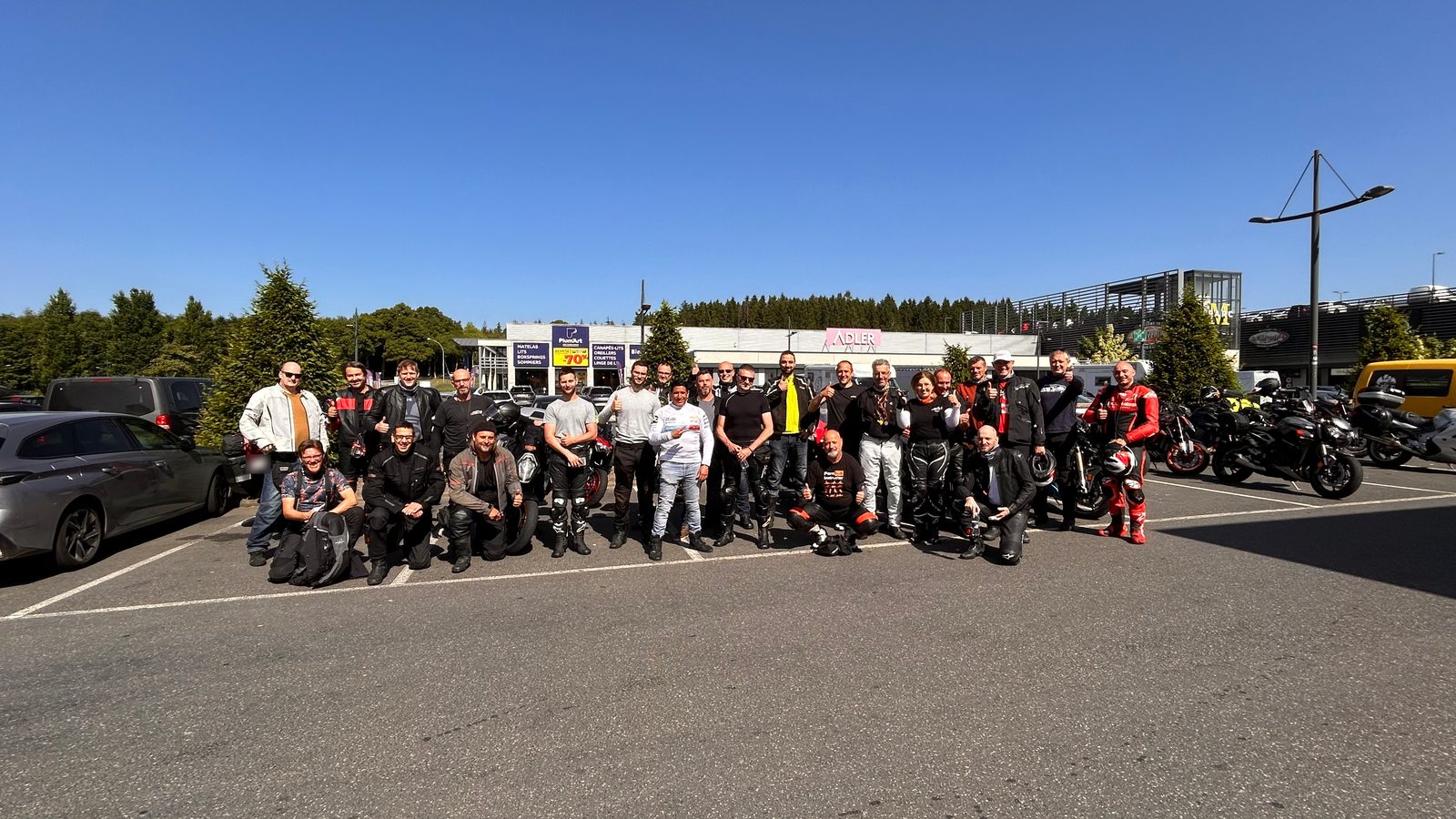 Een grote groep motorrijders poseert in de zon op een parking, sommigen gehurkt vooraan, velen met de duim omhoog, met hun motoren ernaast.