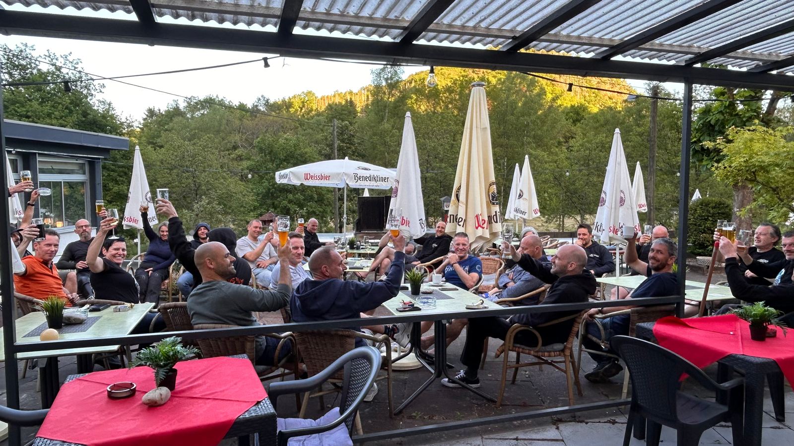 Een grote groep heft de glazen op een overdekt terras met parasols en bos op de achtergrond.