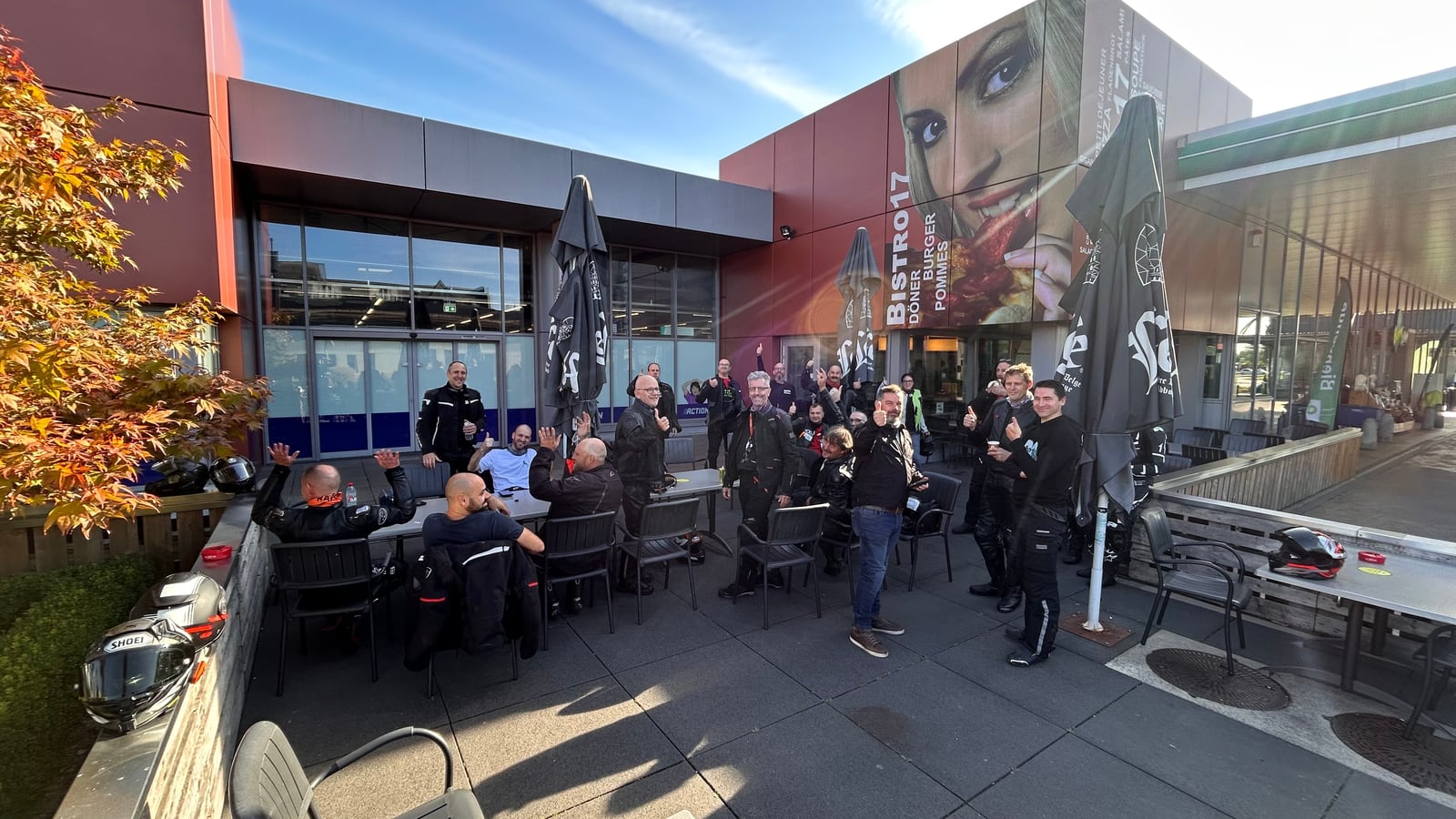 Een groep motorrijders lacht en zwaait op een terras tijdens een koffiestop.