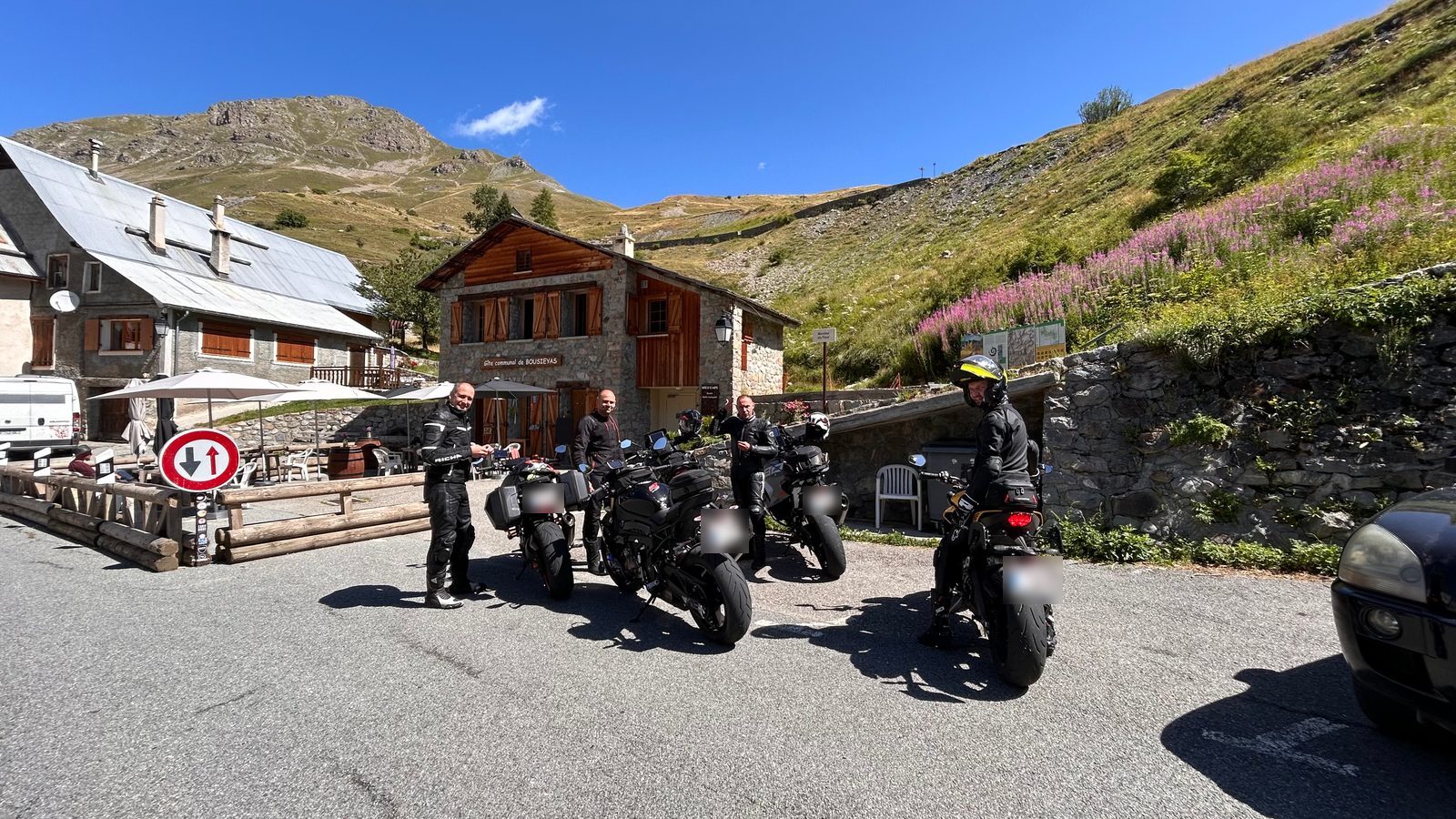 Vier motorrijders bij hun motoren voor een stenen berghut met terras, met groene bergflanken en een strakblauwe lucht.