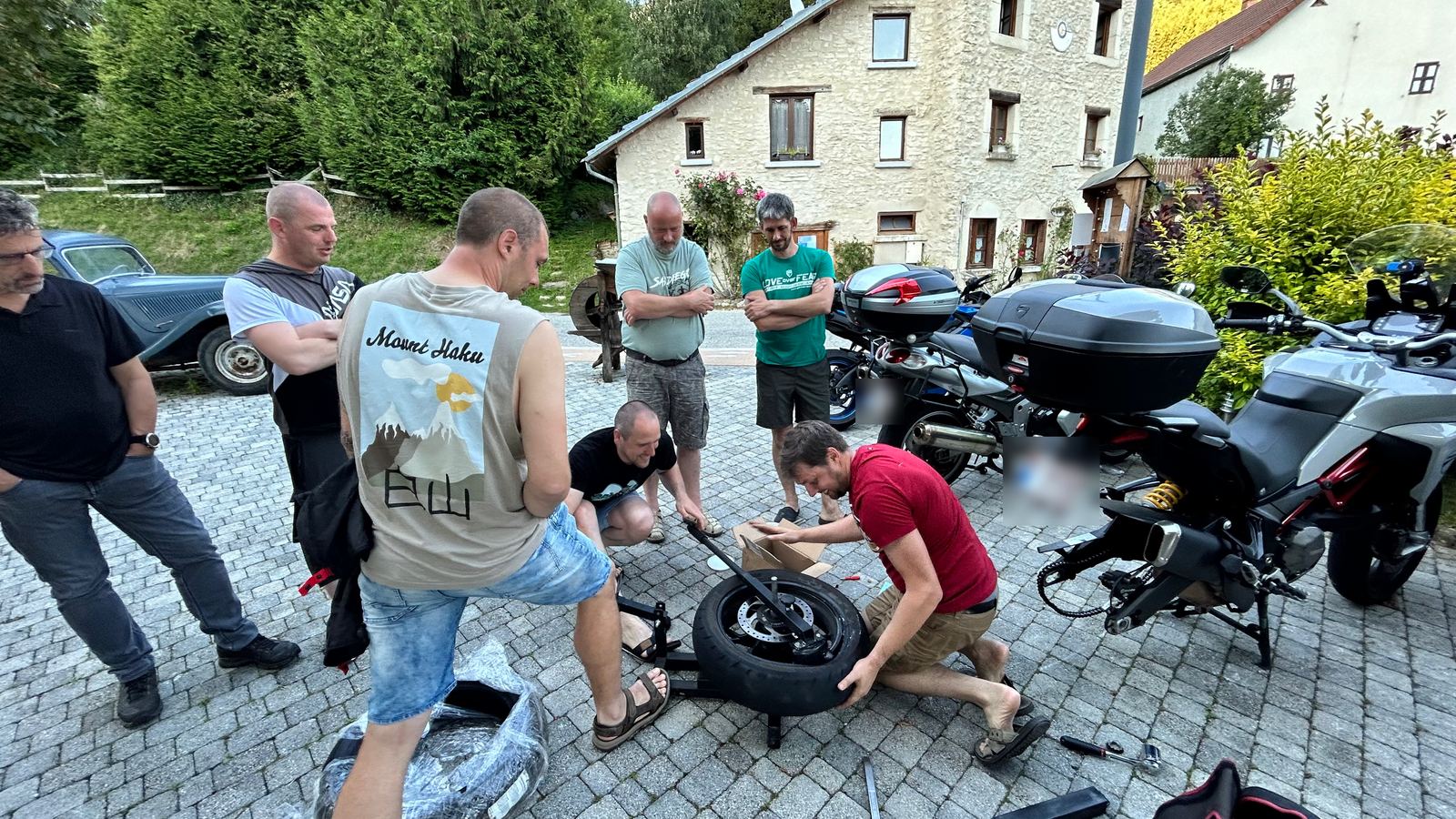 Een groep kijkt toe hoe twee motorrijders samen een motorband wisselen op een binnenplein.