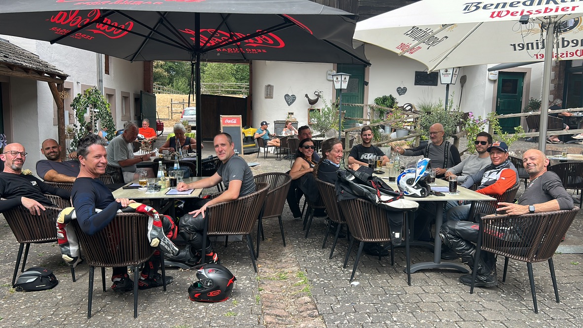 Motorrijders zitten samen aan tafels op een terras, met helmen naast zich.