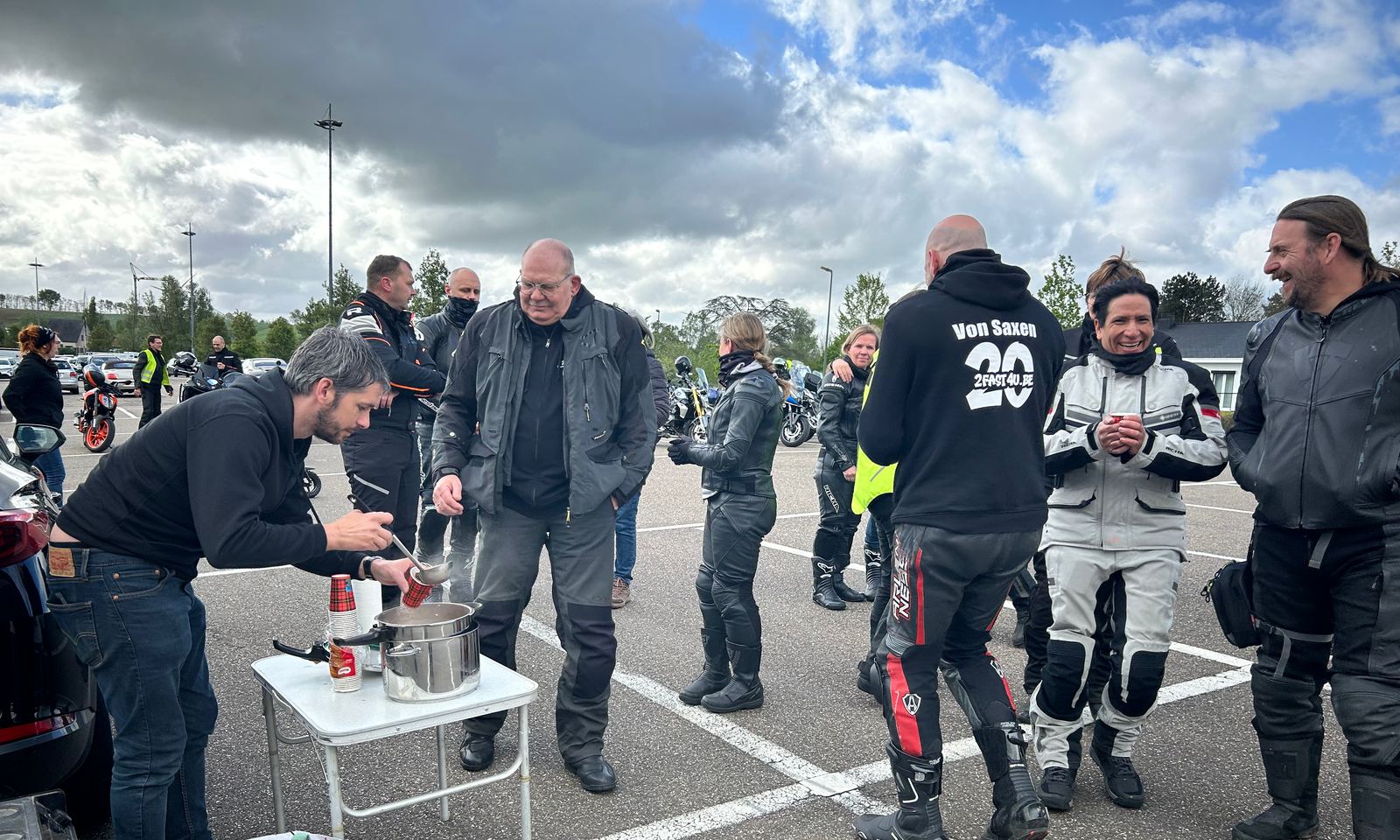 Motorrijders staan lachend en pratend op een parking terwijl iemand soep uitschept aan een klaptafel.