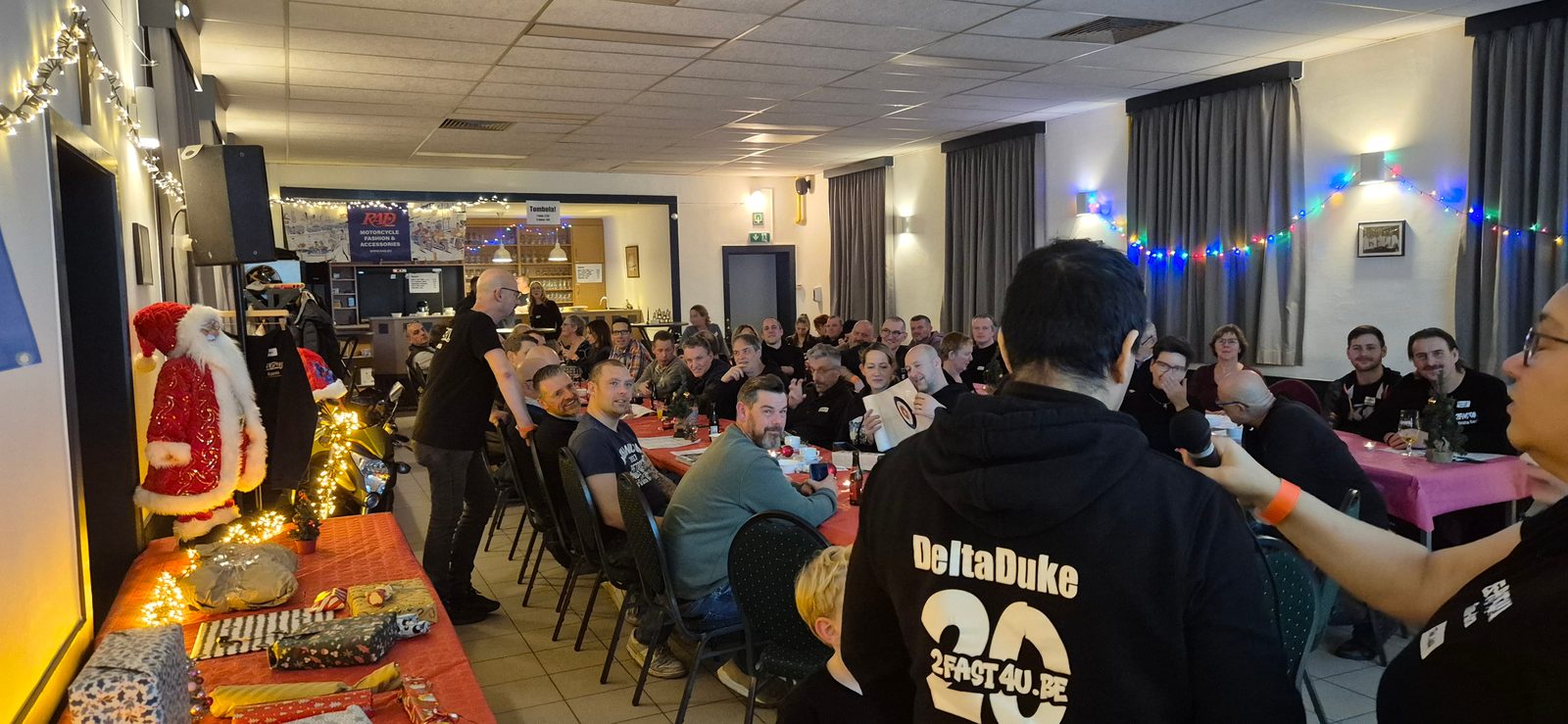 Een volle zaal op de WinterBBQ van november 2025, met links een kerstman en een tafel vol ingepakte cadeautjes en vooraan iemand in een 2fast4u-hoodie die de microfoon krijgt.