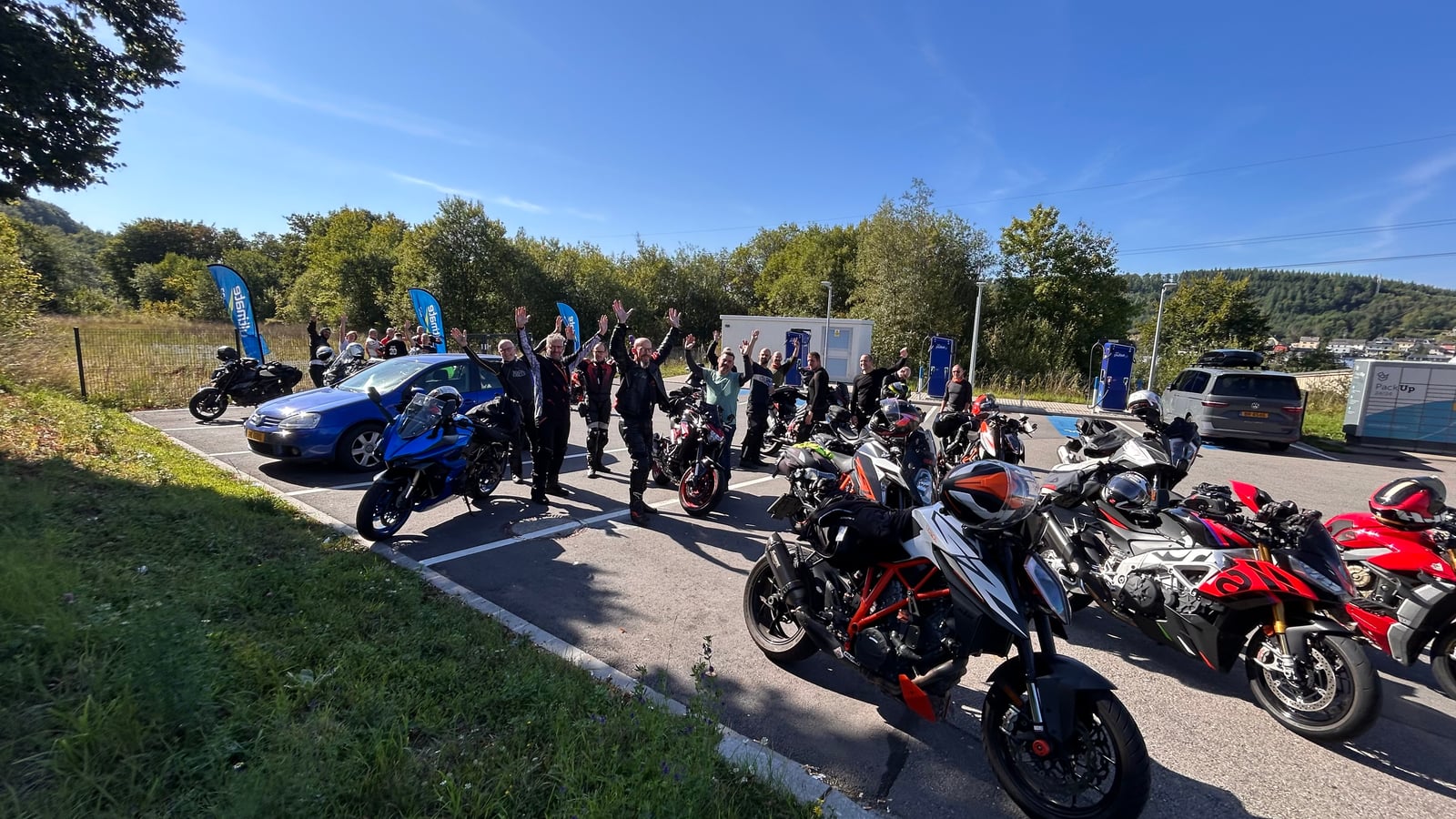 Motorrijders zwaaien naar de camera naast een rij motoren op een zonnige parkeerplaats.